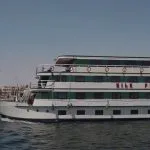 MS Nile Plaza cruise ship sailing on the Nile River under a clear blue sky, showing its white and green exterior.