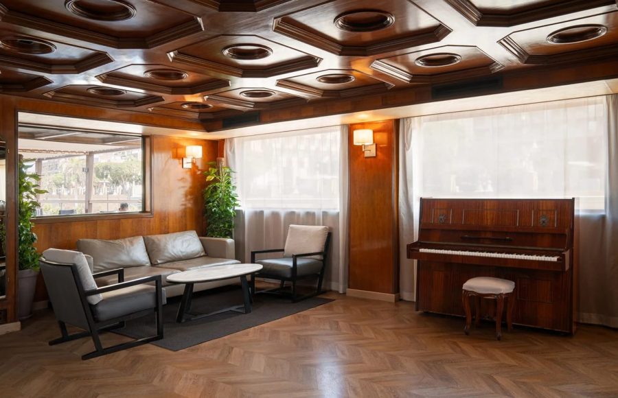 Elegant lounge on the MS Nile Plaza featuring a glossy wooden coffered ceiling, a piano, modern seating, and large windows.