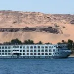 A large white multi-deck Nile cruise ship sailing on the river with desert hills and ancient tombs in the background.