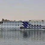 A full-side exterior view of the M/S Radamis II, a white and blue multi-deck Nile cruise ship, sailing on the river under a clear sky with palm trees and green banks visible in the background.