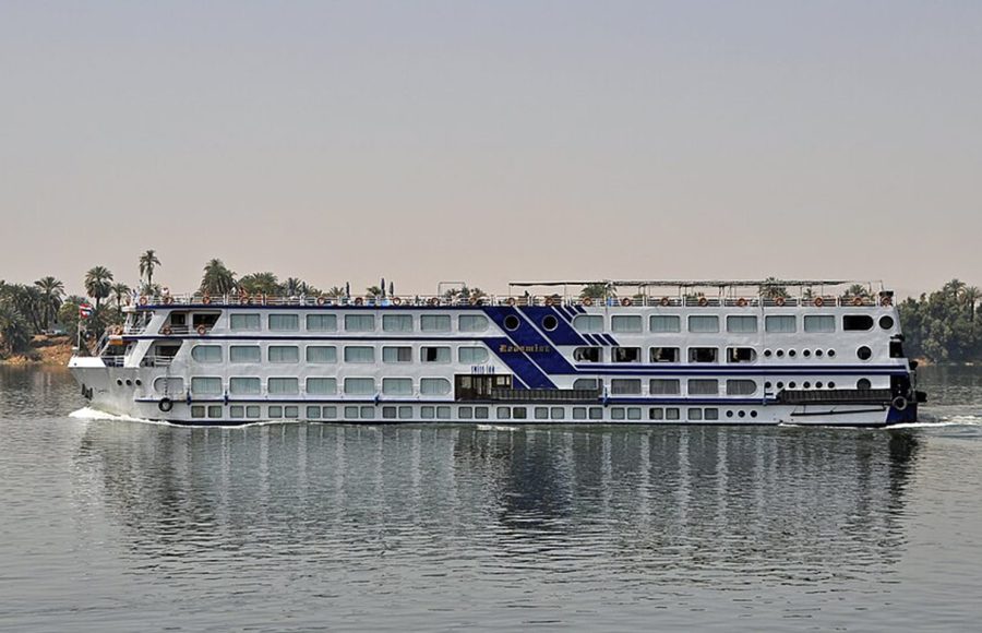 A full-side exterior view of the M/S Radamis II, a white and blue multi-deck Nile cruise ship, sailing on the river under a clear sky with palm trees and green banks visible in the background.