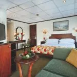 A spacious Nile cruise ship cabin featuring a double bed with a colorful floral throw, a green chaise lounge, a wooden TV cabinet and flat-screen TV, a desk/vanity area with a mirror, and a large window providing a view of the river.