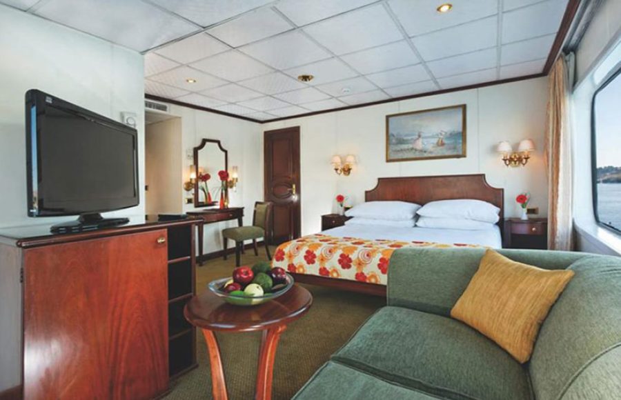 A spacious Nile cruise ship cabin featuring a double bed with a colorful floral throw, a green chaise lounge, a wooden TV cabinet and flat-screen TV, a desk/vanity area with a mirror, and a large window providing a view of the river.