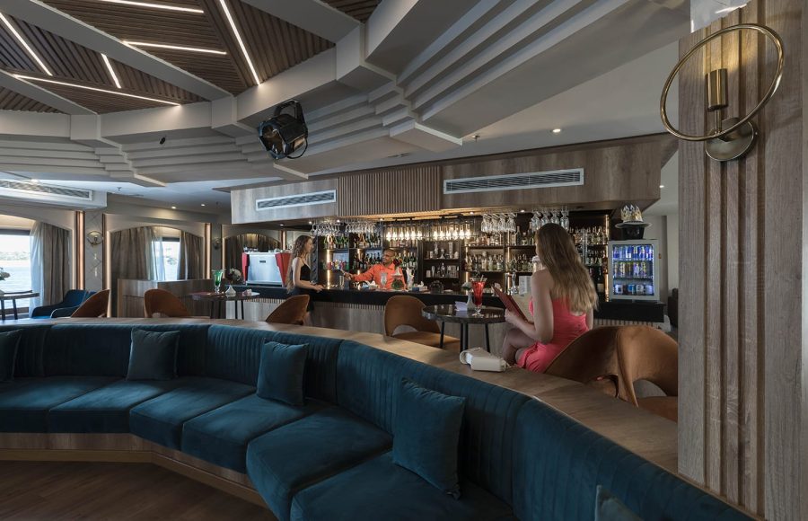 Modern, well-lit cruise ship bar with a large, curved teal sofa in the foreground.