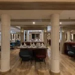 Interior of a ship restaurant with light wood floors, white fluted columns, and dining tables set with dark green and burgundy chairs.