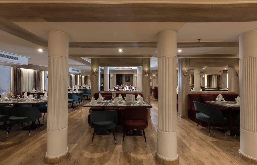 Interior of a ship restaurant with light wood floors, white fluted columns, and dining tables set with dark green and burgundy chairs.