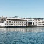 Side view of the white MS Royal Beaurivage Nile cruise ship with multiple decks and rows of windows, sailing on calm blue water under a clear sky.