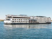 Side view of the white MS Royal Beaurivage Nile cruise ship with multiple decks and rows of windows, sailing on calm blue water under a clear sky.