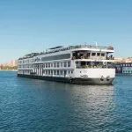 Exterior view of the MS Royal Beaurivage, a white multi-deck Nile cruise ship, sailing on blue water with a city skyline in the background.