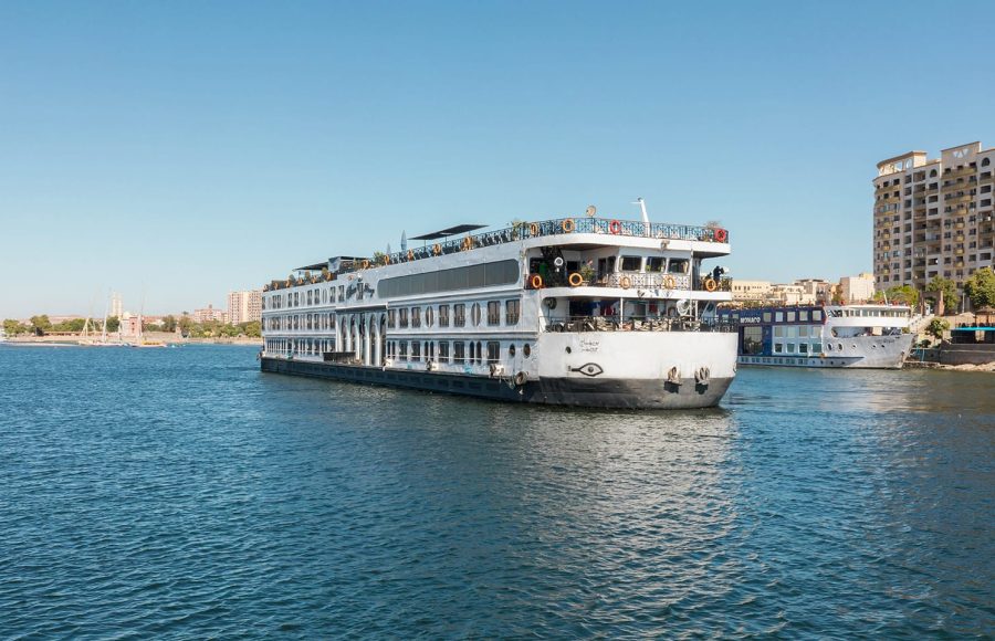 Exterior view of the MS Royal Beaurivage, a white multi-deck Nile cruise ship, sailing on blue water with a city skyline in the background.