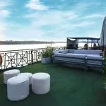 A sunny outdoor sundeck with modern grey lounge furniture, white ottomans, and potted plants, overlooking a large river.