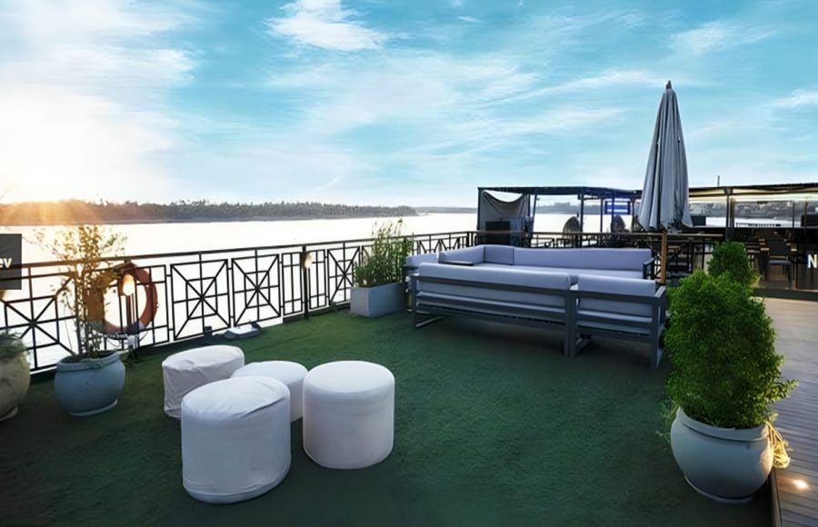A sunny outdoor sundeck with modern grey lounge furniture, white ottomans, and potted plants, overlooking a large river.