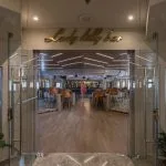View through glass doors into the "Lady Lilly Bar" on a cruise ship, showing a spacious lounge with high tables, stools, and a central bar area.