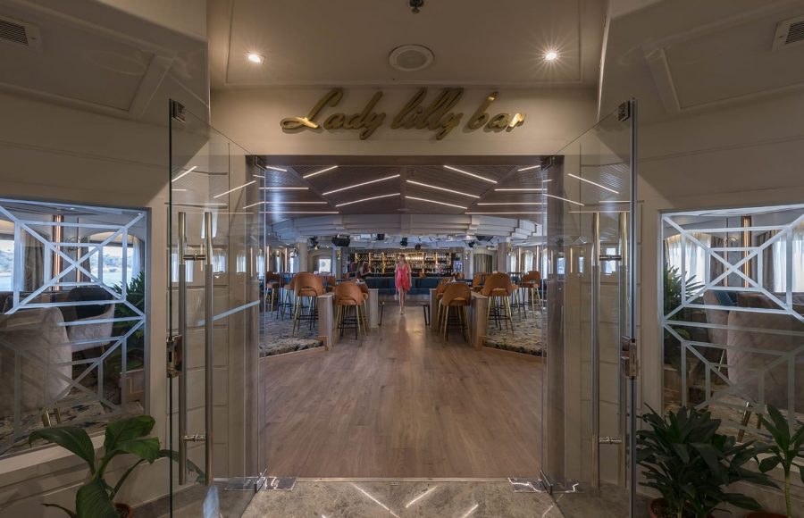 View through glass doors into the "Lady Lilly Bar" on a cruise ship, showing a spacious lounge with high tables, stools, and a central bar area.