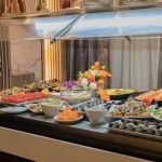 Cold buffet station under a sneeze guard, featuring plates of salads, sliced tomatoes, sushi rolls, and decorative carved vegetables.