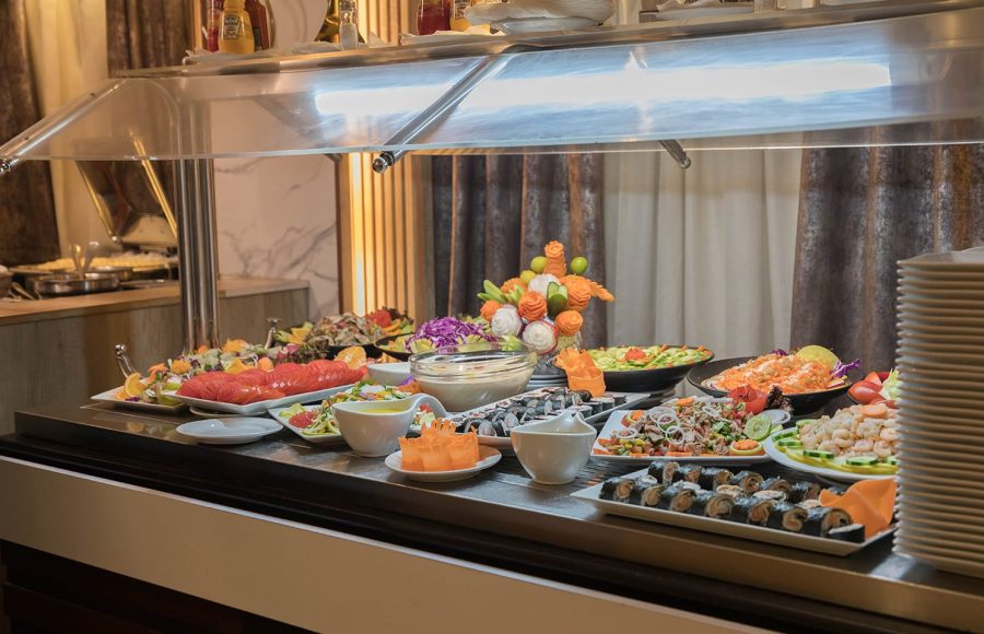 Cold buffet station under a sneeze guard, featuring plates of salads, sliced tomatoes, sushi rolls, and decorative carved vegetables.