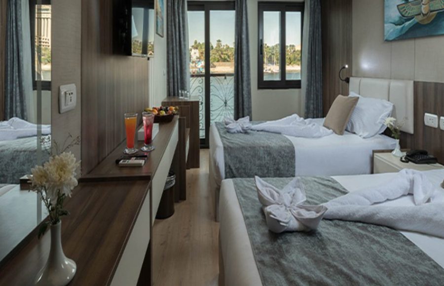 Bright twin-bed cruise cabin with dark wood finishes, decorative towels, and a balcony view of a sunny riverbank.