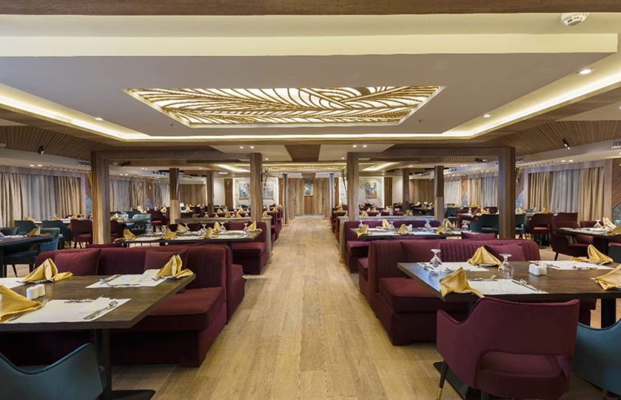 Cruise ship restaurant with dark burgundy banquettes, wooden tables set with gold napkins, and a decorative gold leaf ceiling panel.