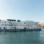 The MS Royal Elite, a large, white, multi-deck river cruise ship, sailing on the Nile River on a sunny day with buildings and sandy hills visible on the banks.