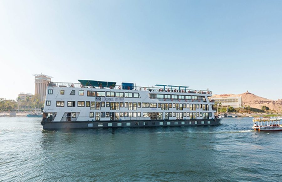 The MS Royal Elite, a large, white, multi-deck river cruise ship, sailing on the Nile River on a sunny day with buildings and sandy hills visible on the banks.