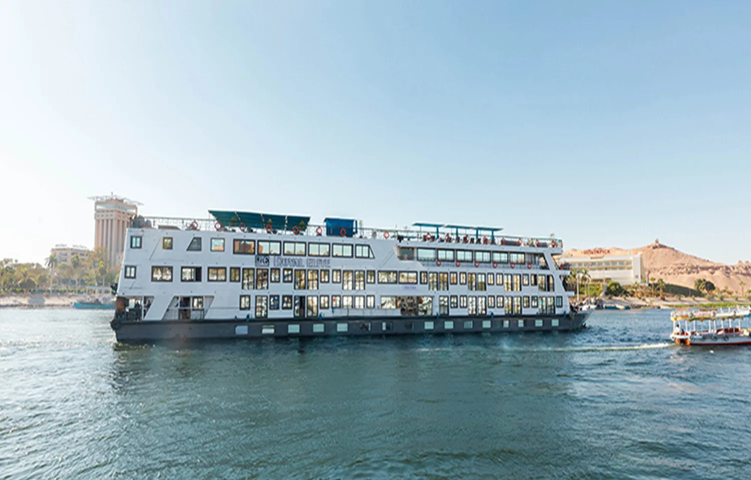 The MS Royal Elite, a large, white, multi-deck river cruise ship, sailing on the Nile River on a sunny day with buildings and sandy hills visible on the banks.