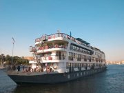 Three-quarter view of the multi-deck MS Royal Elite river cruise ship with life preservers and exterior stairs, sailing on the Nile River.