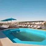 Cruise ship sundeck with a bright blue swimming pool, surrounded by rows of lounge chairs and umbrellas, with a view of the river and land.