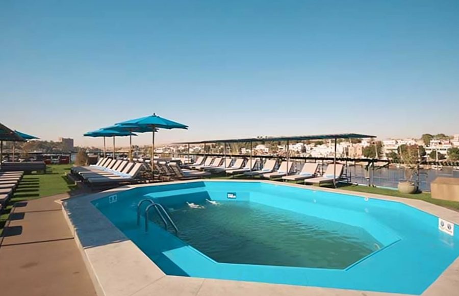 Cruise ship sundeck with a bright blue swimming pool, surrounded by rows of lounge chairs and umbrellas, with a view of the river and land.