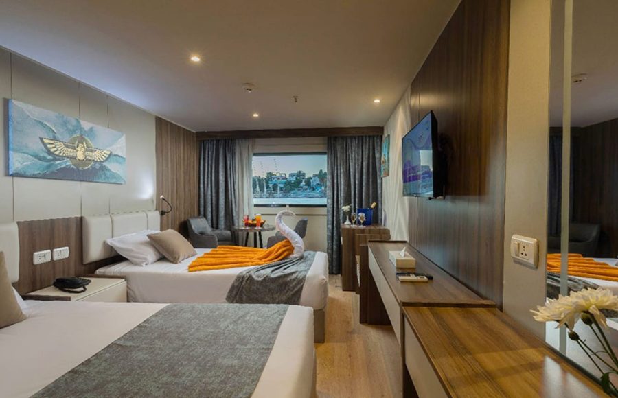 Cruise ship cabin with two twin beds, Egyptian-themed artwork above the headboard, and a towel swan accent on the bed.
