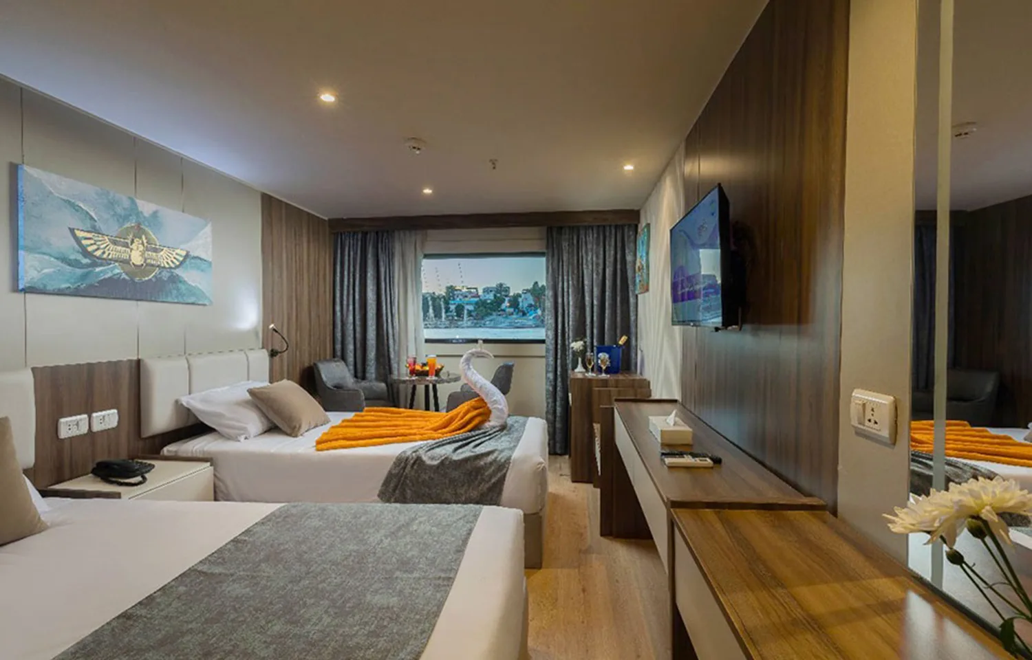 Cruise ship cabin with two twin beds, Egyptian-themed artwork above the headboard, and a towel swan accent on the bed.