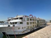 The M/S Royal Viking Nile cruise ship docked alongside a stone quay, showing its multi-deck white and tan exterior, with Arabic text on the bow and passengers visible on the upper decks.