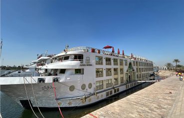 The M/S Royal Viking Nile cruise ship docked alongside a stone quay, showing its multi-deck white and tan exterior, with Arabic text on the bow and passengers visible on the upper decks.