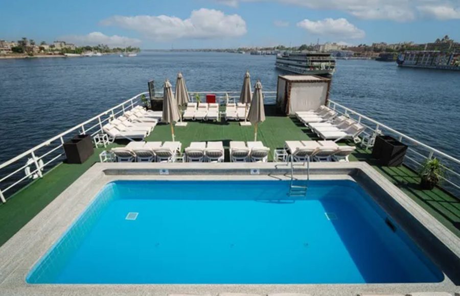 Wide view of a Nile cruise ship's sundeck with a rectangular swimming pool in the foreground and rows of sun loungers and umbrellas overlooking the river.