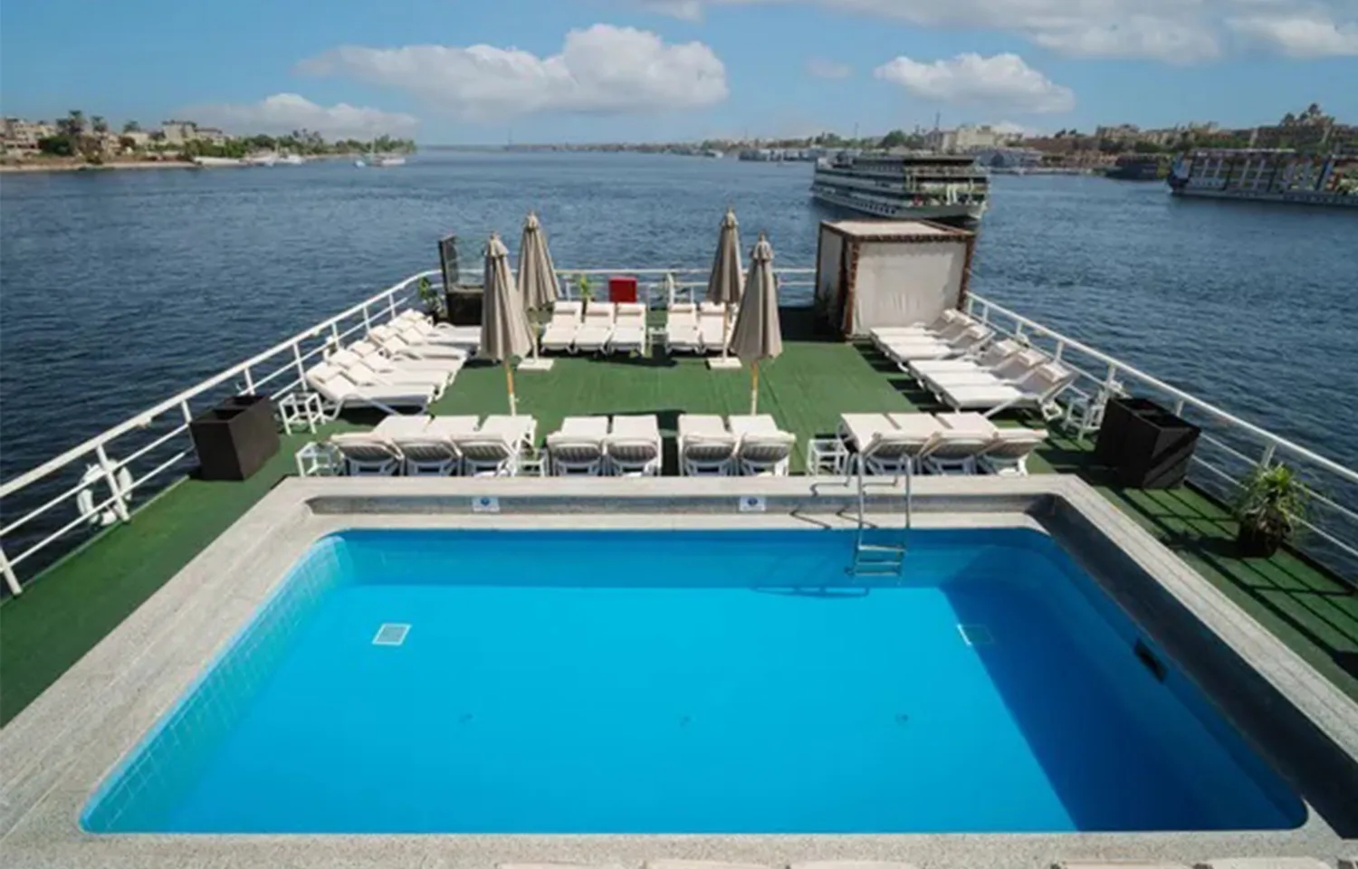Wide view of a Nile cruise ship's sundeck with a rectangular swimming pool in the foreground and rows of sun loungers and umbrellas overlooking the river.