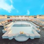 A geometric swimming pool on the wooden sundeck of a river cruise ship, lined with lounge chairs and overlooking a wide river with other docked vessels in the background.