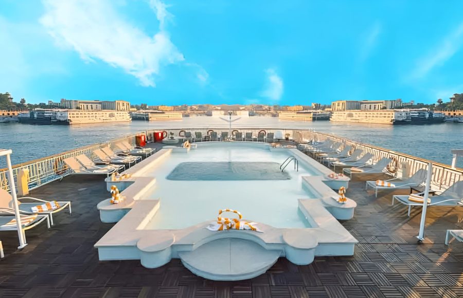 A geometric swimming pool on the wooden sundeck of a river cruise ship, lined with lounge chairs and overlooking a wide river with other docked vessels in the background.