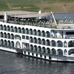 The exterior of a multi-deck white Nile cruise ship with arched windows, a sun deck, and a small pool, sailing on the river.