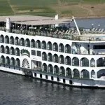 The exterior of a multi-deck white Nile cruise ship with arched windows, a sun deck, and a small pool, sailing on the river.