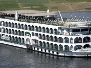 The exterior of a multi-deck white Nile cruise ship with arched windows, a sun deck, and a small pool, sailing on the river.