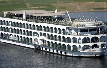 The exterior of a multi-deck white Nile cruise ship with arched windows, a sun deck, and a small pool, sailing on the river.