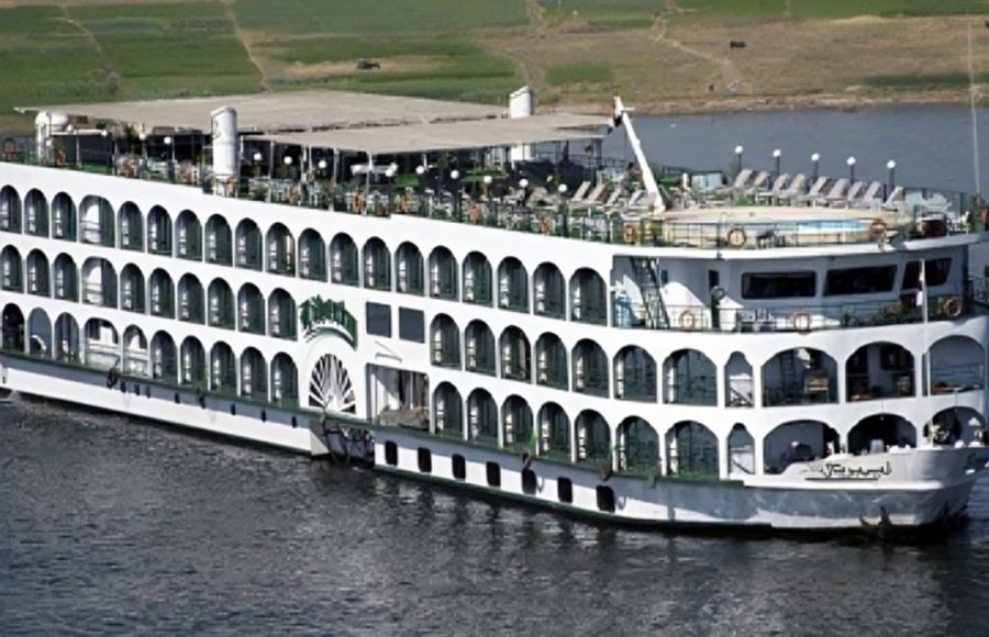 The exterior of a multi-deck white Nile cruise ship with arched windows, a sun deck, and a small pool, sailing on the river.