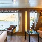 The interior of a luxury suite on the M/S Alyssa Nile cruise ship featuring a seating area with an orange sofa, colorful patterned armchairs, and a large panoramic window overlooking the Nile River and desert hills.