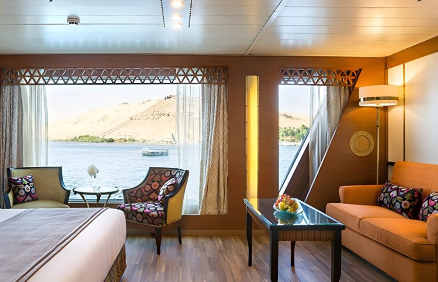 The interior of a luxury suite on the M/S Alyssa Nile cruise ship featuring a seating area with an orange sofa, colorful patterned armchairs, and a large panoramic window overlooking the Nile River and desert hills.