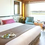 Interior of a bright, modern bedroom in a suite on the M/S Alyssa Nile cruise ship, featuring a king-sized bed with pink geometric pillows and a large window offering a scenic view of the Nile River.