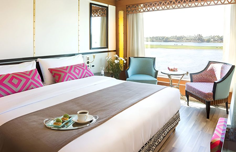 Interior of a bright, modern bedroom in a suite on the M/S Alyssa Nile cruise ship, featuring a king-sized bed with pink geometric pillows and a large window offering a scenic view of the Nile River.