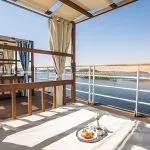A luxury sun deck on the Alyssa Nile cruise ship featuring wooden cabanas with white and blue striped pillows and curtains, overlooking the Nile River and desert hills.