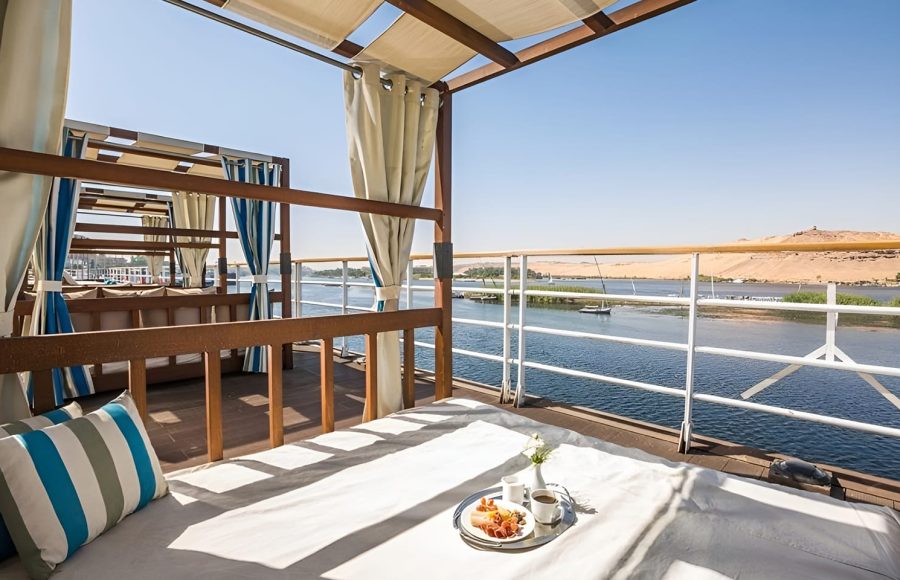 A luxury sun deck on the Alyssa Nile cruise ship featuring wooden cabanas with white and blue striped pillows and curtains, overlooking the Nile River and desert hills.