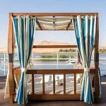 A high-angle view of luxury wooden cabanas on the sun deck of the Alyssa Nile cruise ship, featuring striped curtains and a breakfast tray, with the Nile River and desert hills in the background.