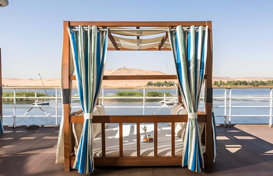 A high-angle view of luxury wooden cabanas on the sun deck of the Alyssa Nile cruise ship, featuring striped curtains and a breakfast tray, with the Nile River and desert hills in the background.