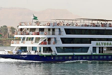 A Multi-Deck Luxury Nile Cruise Ship Named &Amp;Quot;Alyssa&Amp;Quot; Sailing On The River In Egypt, Featuring A Blue And White Hull With Passengers On The Top Deck And Desert Hills In The Background.